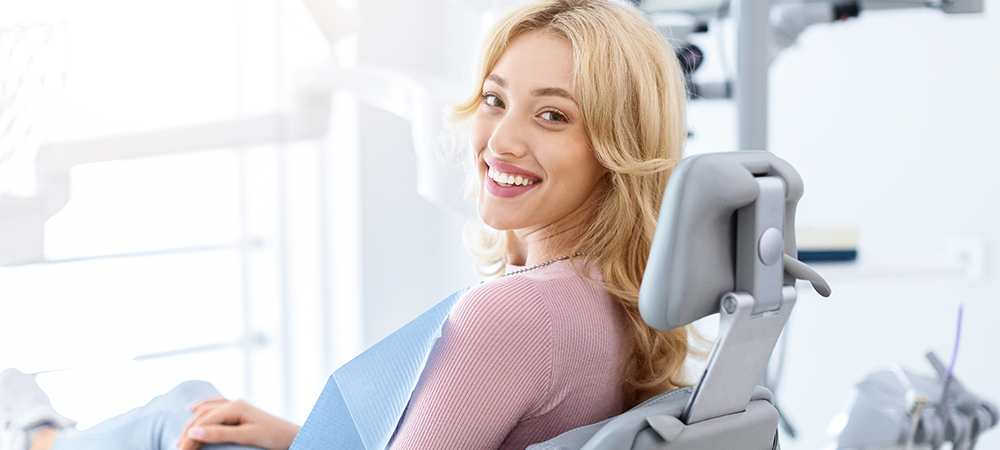 Smiling and relaxed young woman sitting at dental chair in modern clinic, waiting for her dentist. Happy attractive blonde lady attending luxury dental clinic