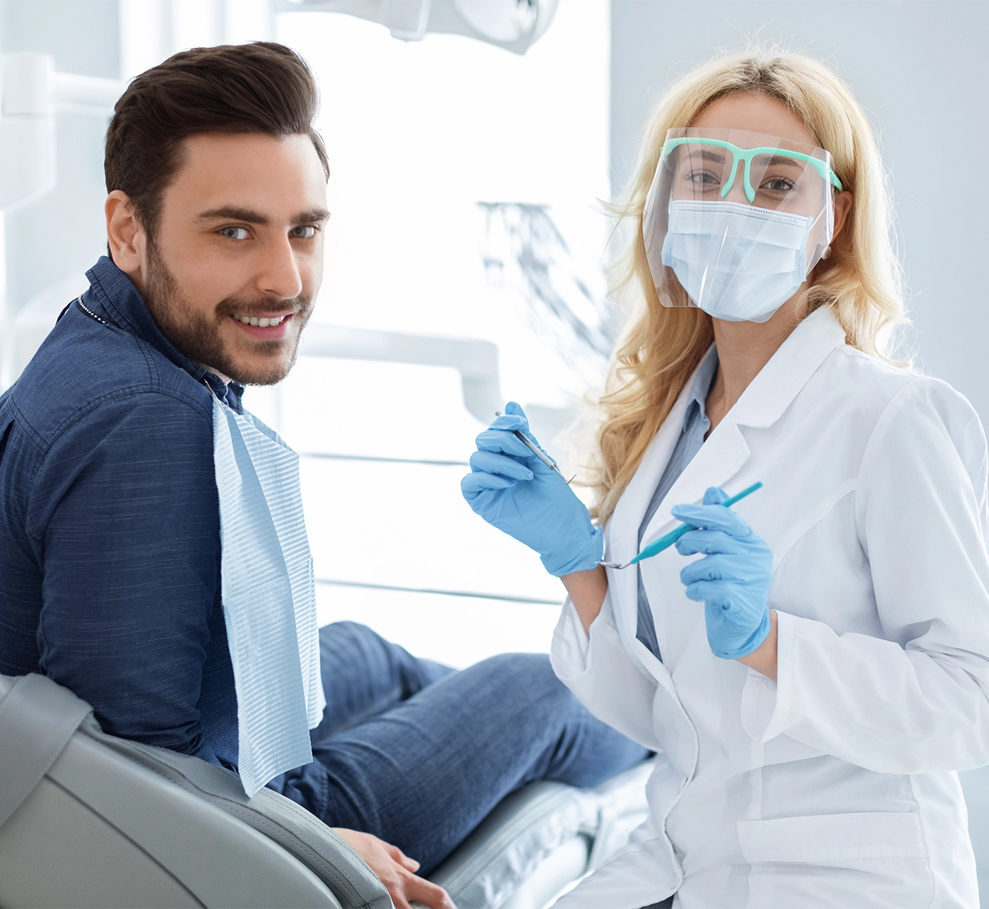 Dental clinic patient happy young bearded man and female doctor in workwear holding dental tools, looking together at camera, dentist making check up for cheerful guy, modern dental cabinet interior