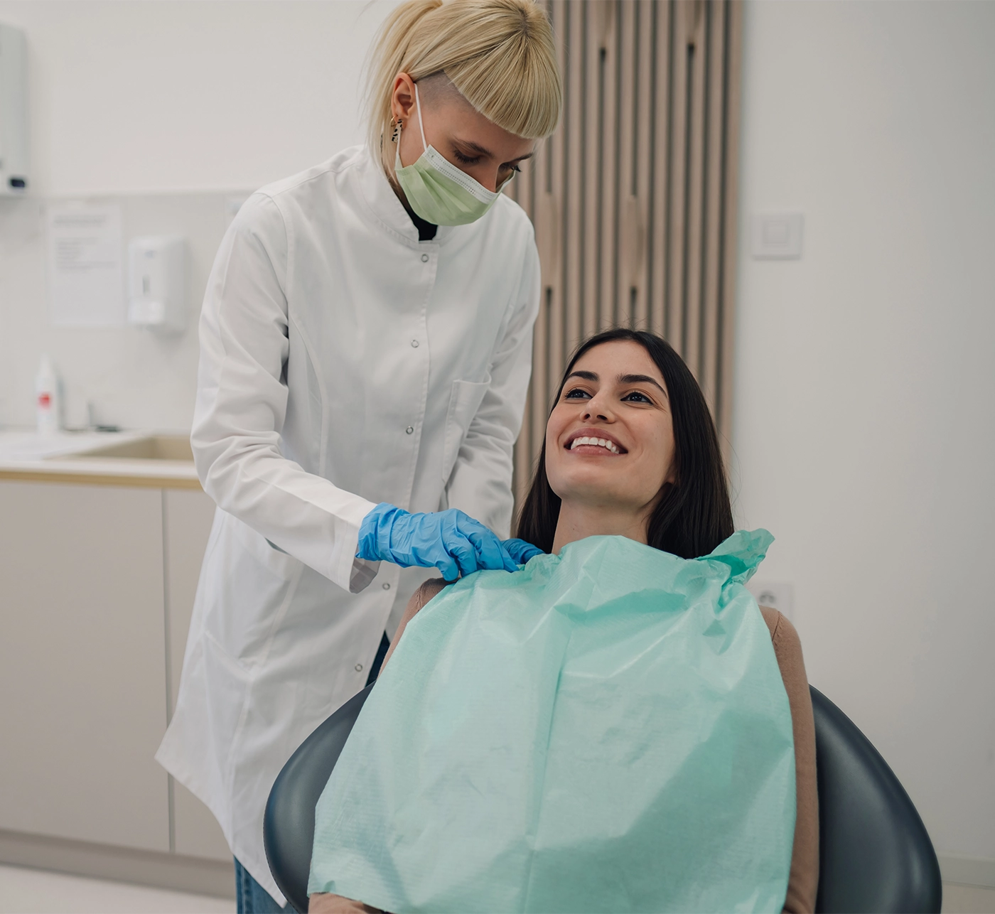 Female dentist wearing mask and gloves preparing smiling female patient for dental checkup in modern dental clinic