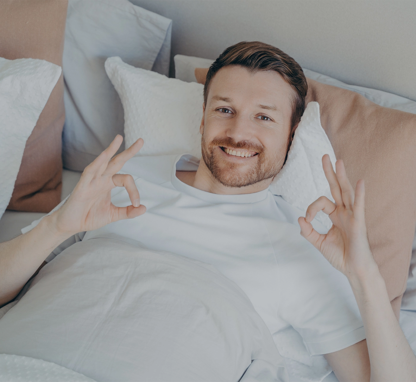 Relaxed young bearded man lying in his bed after waking up in early morning, enjoying leisure time while staying at home on weekend, satisfied guy showing ok sign with both hands